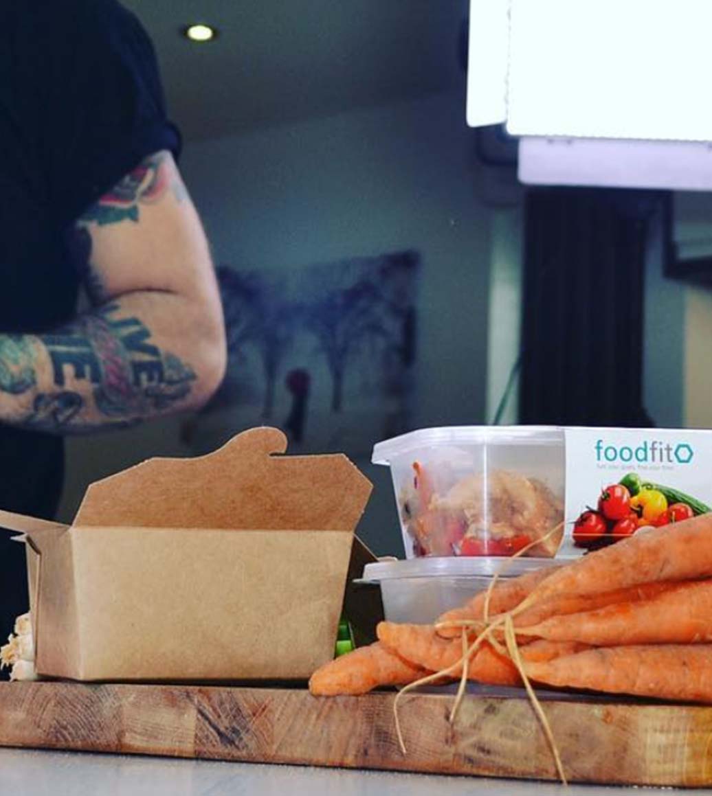 A kitchen countertop with fresh vegetables and Food Fit subscription box shows you can learn more about the Food Fit kitchen.