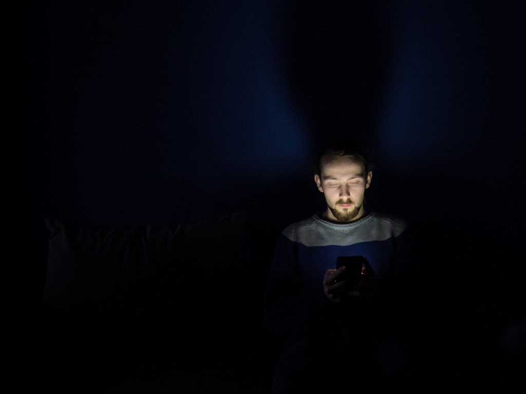 Man with a beard, holding a phone in the dark, implies the effects of blue light on sleep.